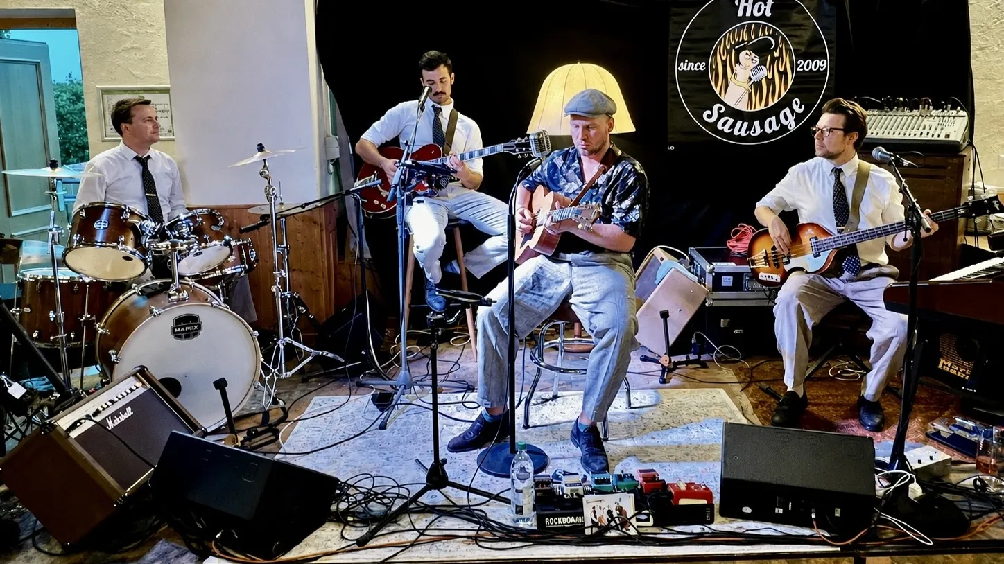Hot Sausage spielt sitzend ein akustisches Set vor dem Bandbanner